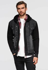 Giacca in denim nera e grigia con cappuccio, dotata di tasche frontali e chiusura a bottoni, indossata sopra una camicia a quadri rossa e bianca, abbinata a jeans neri.