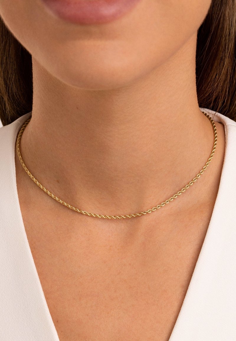 Gros plan sur le cou d'une femme portant un collier fin en chaîne d'or torsadée et un haut blanc.