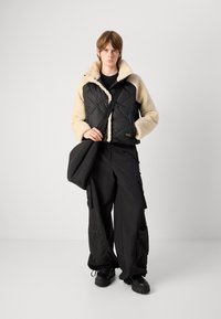 Μαύρο επενδυμένο μπουφάν με κρεμ λεπτομέρειες από γούνα κ�αι ψηλό γιακά, συνδυασμένο με φαρδιά μαύρα cargo παντελόνια και μαύρα χοντρά παπούτσια.