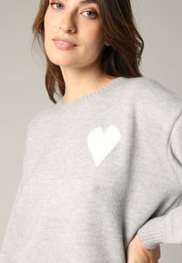 Pull en tricot gris clair avec un col rond, présentant un motif de cœur texturé blanc sur la poitrine gauche et des manches côtelées.