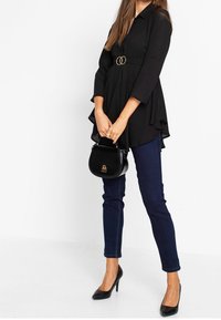 Blouse noire avec un design superposé, bagues dorées à la taille, associée à un jean skinny bleu foncé et des escarpins noirs à bout pointu, tenant un sac à main noir.
