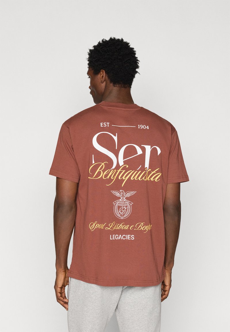 Καφέ βαμβακερό μπλουζάκι με άνετη εφαρμογή. Διαθέτει μεγάλα λευκά και κίτρινα γράμματα, σχέδιο εμβλήματος και κοντά μανίκια για μια χαλαρή εμφάνιση.