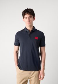 Marineblå polo skjorte i pustende materiale med tre knapper. Har et fremtredende rødt rektangulært logo-merke på brystet.