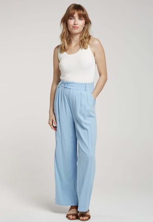 Pantalons larges bleu clair avec des plis, présentant une taille haute et des détails de bouton. Assortis à un débardeur blanc ajusté et des sandales marron.