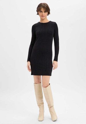 Kobieta w dopasowanej, czarnej sukience mini z długim rękawem, ozdobionej subtelnym pionowym wzorem, oraz w beżowych kozaczkach do kolan, stojąca na tle jednolitego tła.