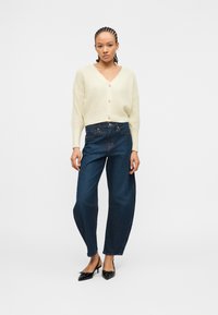 Femme debout portant un cardigan crème boutonné, un jean large foncé et des escarpins noirs à bout pointu, sur un fond blanc uni.