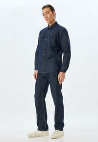 Mörk denimskjorta med två bröstfickor, knäppning och lång ärm, i kombination med mörka denimbyxor och ljusa skor.