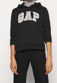 Sweat à capuche noir avec une poche kangourou à l'avant, un lettrage "GAP" imprimé en léopard blanc, et des manches avec poignets. Le tissu est doux, avec une coupe décontractée.