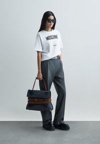 Žena v bielom tričku Moschino, sivých nohaviciach, čiernych slnečných okuliaroch a platformových topánkach, držiaca veľkú čiernu a hnedú kabelku.