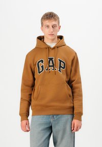 Jeune homme portant un sweat à capuche marron de GAP et un jean bleu clair, se tenant face à un fond blanc uniforme.