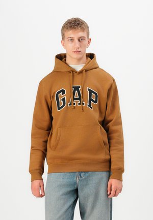 Jeune homme portant un sweat à capuche marron de GAP et un jean bleu clair, se tenant face à un fond blanc uniforme.