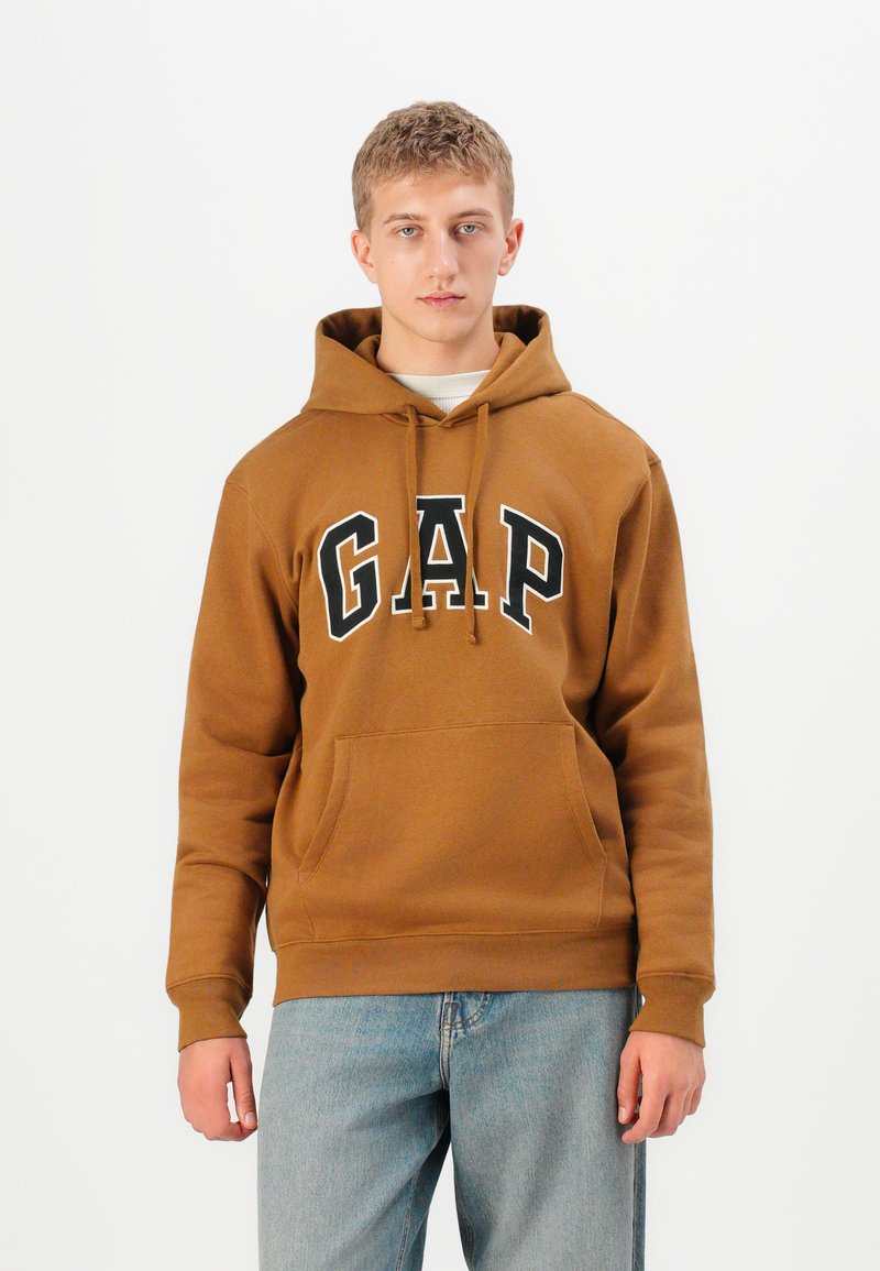 Jeune homme portant un sweat à capuche marron de GAP et un jean bleu clair, se tenant face à un fond blanc uniforme.
