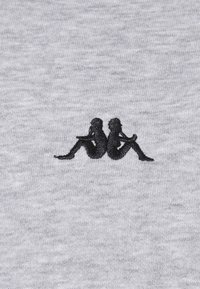 Broderie en silhouette noire de deux femmes assises, dos à dos, sur un fond de tissu gris.
