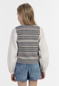 Dziewiarska kamizelka z wielokolorowymi wzorami geometrycznymi, w granatowym i kremowym kolorze, zestawiona z białą, fakturowaną bluzką z długim rękawem oraz dżinsowymi szortami.