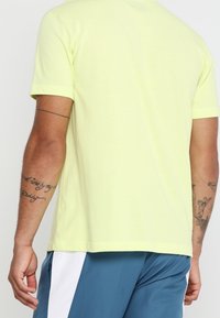 Světle žluté tričko s krátkým rukávem a kulatým výstřihem, které má hladkou texturu. Nošeno s modrými kraťasy, které mají bílé boční detaily.