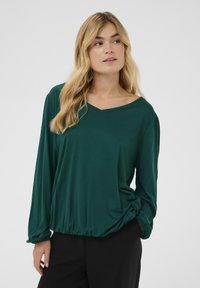 Zeleni top dugih rukava s V-izrezom i skupljenim strukom. Izrađen od mekane tkanine glatke teksture, s lagano širim rukavima.