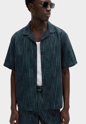 Mann trägt dunkelblaues kurzärmliges Nadelstreifenhemd und passende Hose, weißes Netzunterhemd, schwarzen Gürtel und schwarze Sonnenbrille, seitlich stehend.