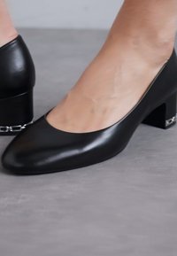 Sorte lærblokk-heels med spiss tå og en subtil kjedeakcent på hælen, med en glatt, blank tekstur.