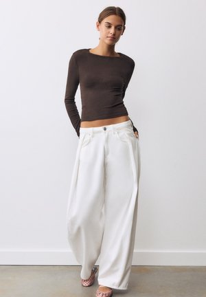 Junge Frau trägt ein enganliegendes dunkles Langarmshirt, weit geschnittene weiße Hosen und rosa Schuhe und steht vor einer schlichten weißen Wand.