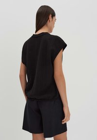 Schwarzes gestricktes Oberteil mit kurzen, weiten Ärmeln und einem lockeren Schnitt. Die Rückseite hat ein leicht strukturiertes Muster, kombiniert mit dunklen Shorts.