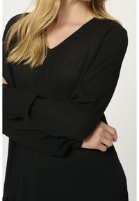Femme aux longs cheveux blonds portant un chemisier noir texturé à col en V et manches longues, les bras croisés.