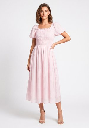 Frau mit welligem braunem Haar, die ein hellrosa Midikleid mit Puffärmeln und beigen Knöchelriemenpumps trägt und mit einer Hand in der Hüfte posiert.