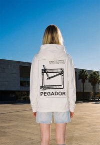 Ξανθό άτομο που φοράει λευκό φούτερ με ένα μαύρο γραφικό καρέκλας και το κείμενο «PEGADOR», στέκεται έξω κοντά σε ένα κτήριο και φοινικόδεντρα.