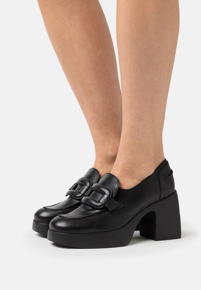 Mocassins à plateforme en cuir noir avec talons carrés et grandes boucles, portés sur des jambes nues sur un fond blanc uni.