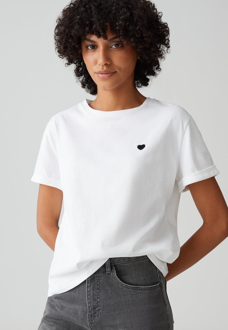 Weißes Baumwoll-T-Shirt mit kurzen, umgeschlagenen Ärmeln, das ein kleines schwarzes Herzmotiv auf der Brust aufweist. Kombiniert mit dunkelgrauen Jeans.