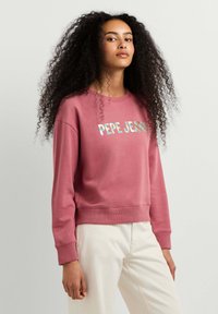 Joven con el pelo largo y rizado, vistiendo una sudadera rosa con el logo "PEPE JEANS" y pantalones claros, de pie frente a un fondo liso.