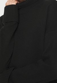 Persona che indossa un maglione a coste nero con maniche lunghe e scollatura alta, mano appoggiata vicino al viso.