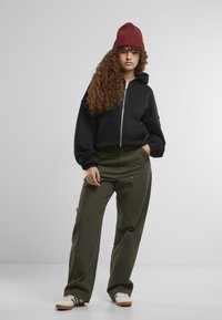 Schwarze Zip-Up-Trainingsjacke, olivgrüne Hose mit seitlichen Knöpfen, elfenbeinfarbene Schuhe mit grünen Streifen und eine gerippte burgunderrote Mütze.
