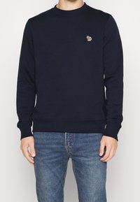 Uomo che indossa una felpa blu scuro con un piccolo logo di zebra multicolore ricamato sul lato sinistro del petto, abbinata a jeans in denim blu.