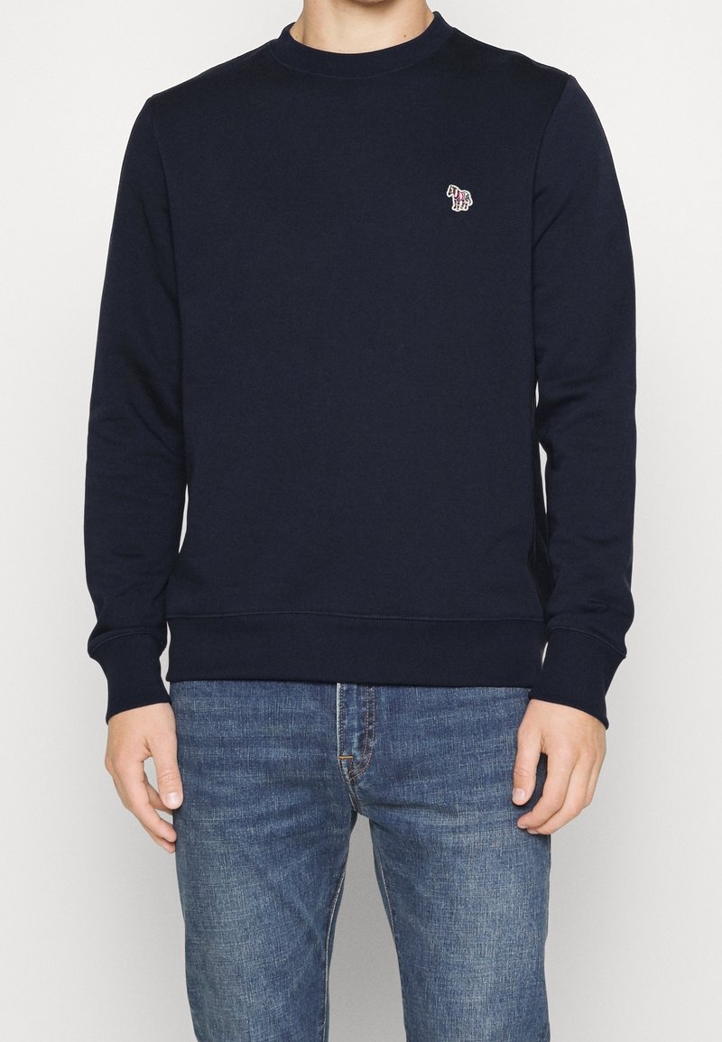 Uomo che indossa una felpa blu scuro con un piccolo logo di zebra multicolore ricamato sul lato sinistro del petto, abbinata a jeans in denim blu.