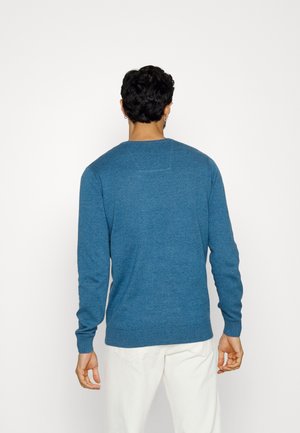 Homme vu de dos portant un pull bleu à manches longues et un pantalon écru sur un fond clair uni.
