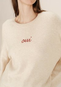 Maglione color crema realizzato in morbido tessuto a maglia. Presenta un ampio scollo e il testo ricamato in rosso "oui" sulla parte frontale.