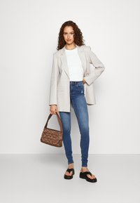 Blazer oversized bege claro, top branco justo, jeans skinny azuis, chinelos pretos e uma mala texturada castanha. Design simples e contemporâneo.