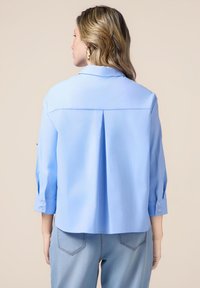 Himmelblaues Hemd mit einer Rückenfalte, Dreiviertelärmeln und knöpfbaren Manschetten. Der Stoff wirkt glatt und hat einen knackigen Kragen.