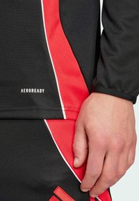 Primer plano de la mano de una persona apoyada sobre ropa deportiva negra y roja con ribetes blancos y el texto "AEROREADY".