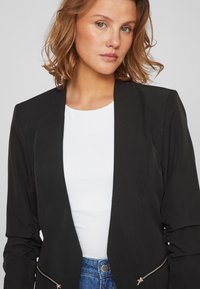 Blazer preto com um design ajustado, apresentando um frente aberta, mangas longas e bolsos com fecho. Usado sobre uma blusa branca e jeans azuis.