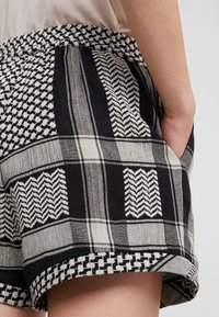 Detailní záběr na krátké kalhoty s černobílým vzorem, pletené, s elastickým pasem a rukou v boční kapse na neutrálním pozadí.