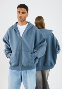 Lichtblauwe zip-up hoodie met een grote capuchon, kangoeroezakken en verlaagde schouders, gecombineerd met een lichte spijkerbroek en grijze broek.
