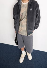 Person som bär en grå Adidas-hoodie, kamouflagemönstrad skjorta, grå shorts, vita strumpor, krämfärgade Adidas-sneakers och håller en liten svart väska.