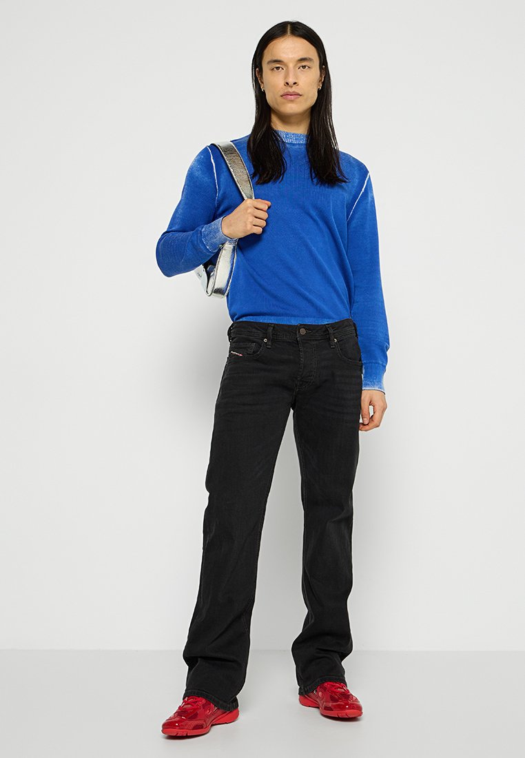 Un pull bleu à manches longues, associé à un jean noir. L'individu porte des baskets rouges brillantes et tient un sac en argent.