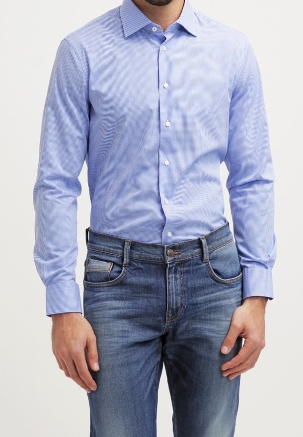 Mand iført en lyseblå ternet skjorte med knapper, stoppet ned i falmede blå jeans, stående med hænderne afslappet ved siden.