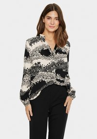Blouse noire et crème avec un motif floral, des manches longues, un devant boutonné et une coupe décontractée. Tissu lisse avec un drapé doux.