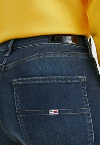 Vaqueros azul oscuro con un bolsillo visible, que presentan costuras en contraste y una pequeña etiqueta de marca. La tela parece suave con una ligera textura.