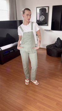 Personne debout les mains dans les poches, portant une salopette vert clair, un t-shirt blanc et des sandales blanches dans un salon avec un sol en bois.