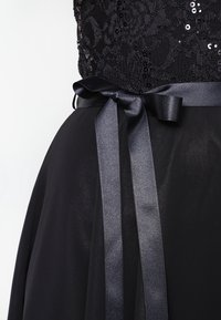 Černé šaty s korzetem z flitrové krajky a splývavou sukní. Saténová stužka zvýrazňuje pas. Hladká textura a elegantní design.