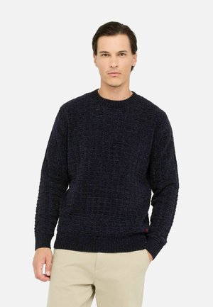 Granatowy sweter dziany z teksturowanym wzorem, ściągaczem na dekolcie i mankietach oraz w pasie. Nosi się go na beżowe spodnie.